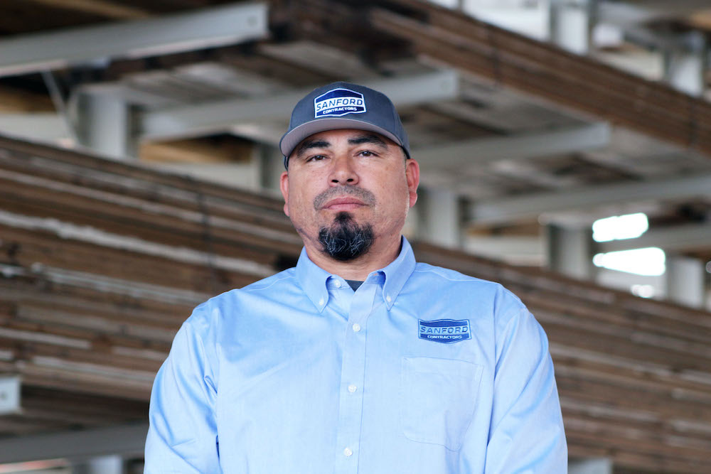 Photo of Luis Sanchez in a warehouse space wearing a Sanford Contractor hat and shirt.