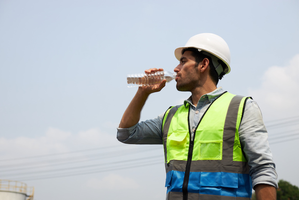 A male construction worker in protective clothing and a helmet, is drinking water to quench his thirst in the harsh sunlight.