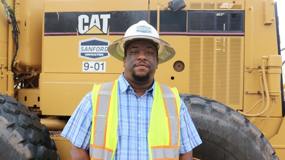Sanford Contractors Site Division Foreman Jay White, wearing a hard helmet, poses with a CAT machine at a construction site.